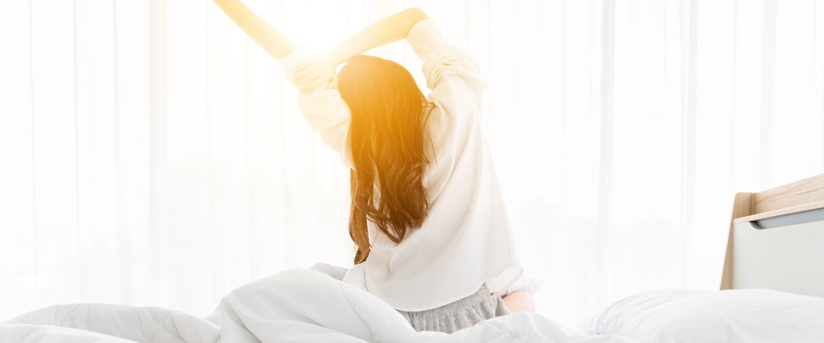 Vrouw rekt zich uit op de rand van haar bed met haar handen in de lucht en de zon schijnt door de gordijnen