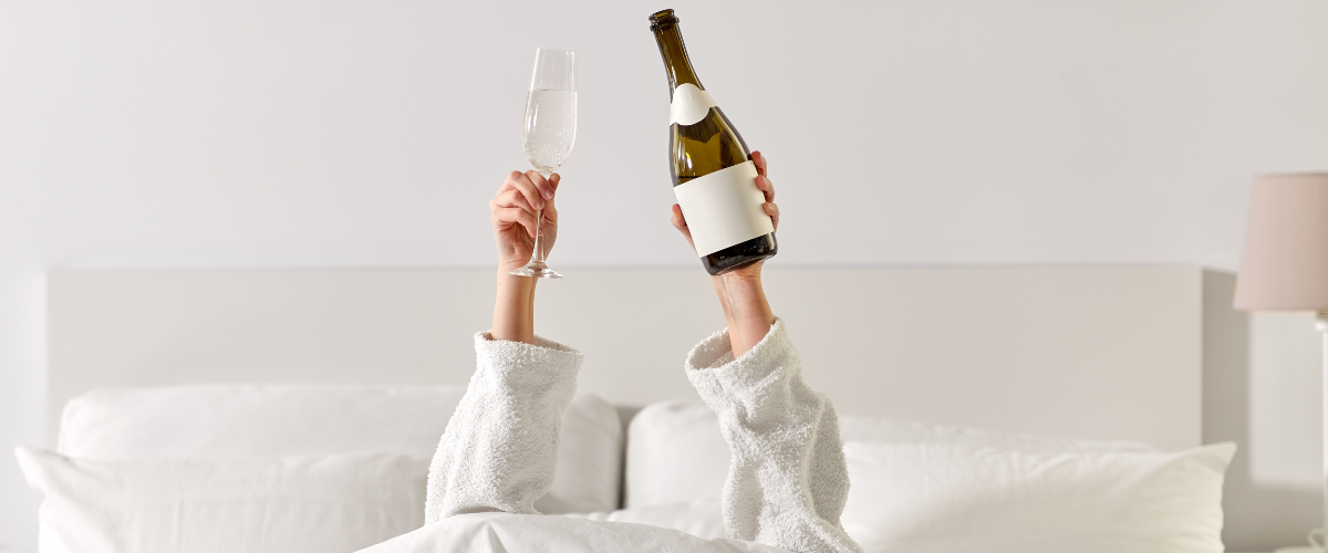 Vrouw ligt in bed en houdt een glas en fles wijn in haar handen omhoog gestoken