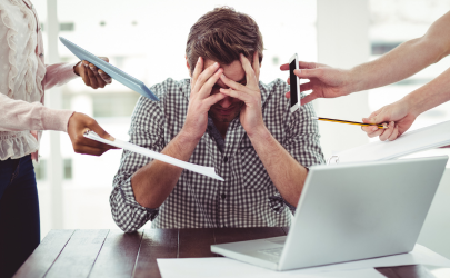 Man zit gefrustreerd aan een bureau met zijn laptop en meerdere mensen om hem heen die iets van hem nodig hebben