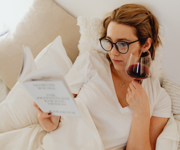 Vrouw ligt in bed een boek te lezen met een glas rode wijn